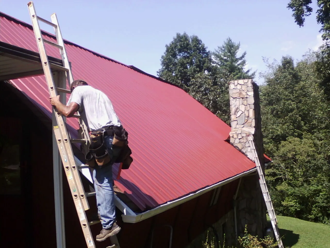 Roofer on a ladder beside a red metal roof and stone chimney — Soffit & Fascia Repair by Roofing Brothers in Monteagle